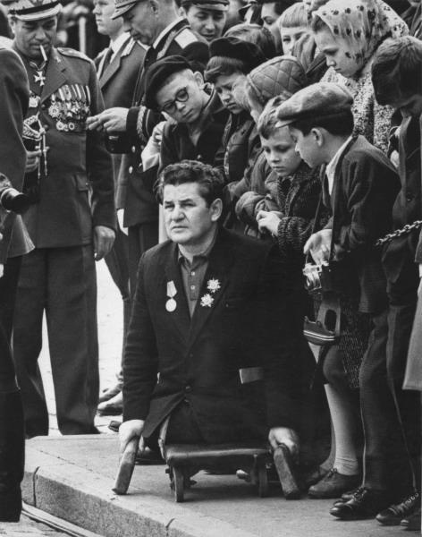 A disabled Soviet veteran with no legs sits in a wheelchair that rolls several inches above the ground. He sits on a street curb surrounded by standing military personnel on his right hand side. A small crowd of children stand to his right and look down at him.