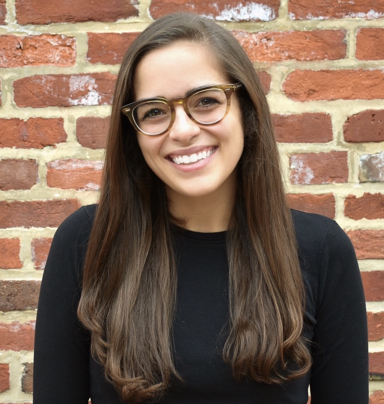 This is a color photograph of a smiling woman in front of a red brick wall. She has long brown hair and is wearing glasses and a black shirt.